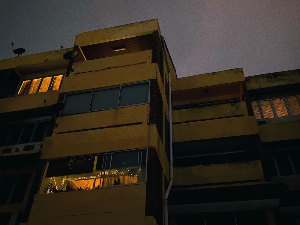 Exterior of old flats, night. Most units unoccupied. 3 have lights on. One of them, ours, is full of plants and fairy lights.